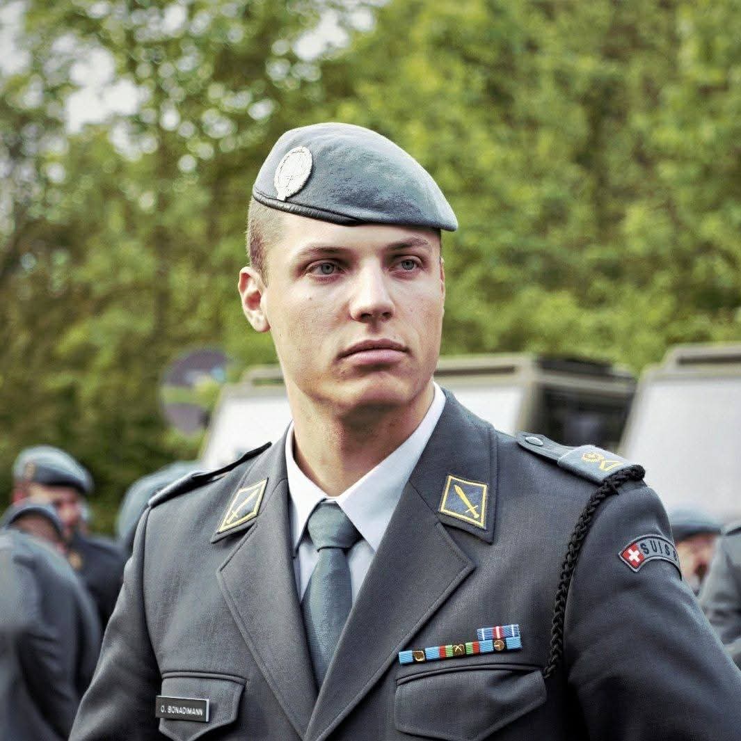 Ein Soldat in Uniform mit Abzeichen steht im Freien, umgeben von Bäumen.